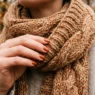 Manicured hand adjusting chunky knit scarf edge in soft autumn light with warm rust tones