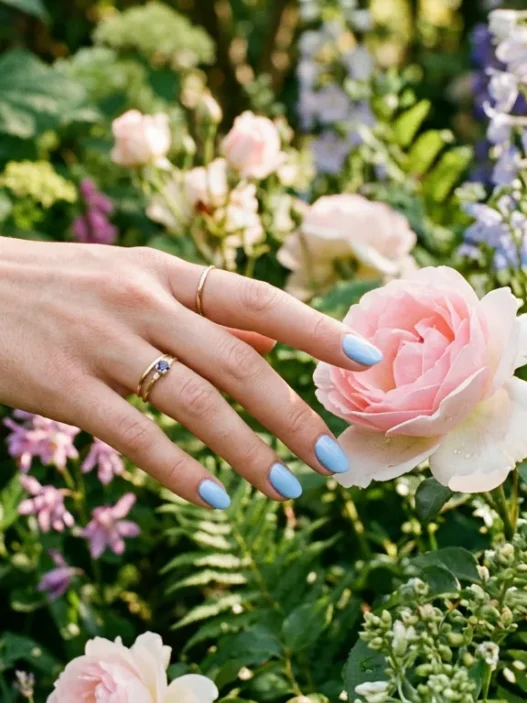 Manicured hand with baby blue nails reaching to touch a single flower bloom in garden setting