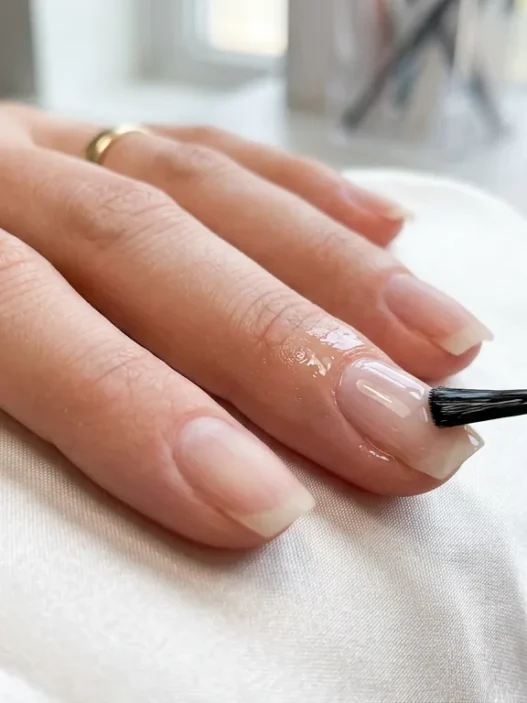 Clear base coat being applied to clean natural nail with brush showing glossy finish