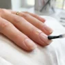 Clear base coat being applied to clean natural nail with brush showing glossy finish