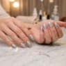 Woman's hands displaying completed holographic foil transfer manicure on marble salon table with mirror finish