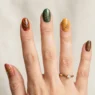 Woman's hand showing various fall nail designs in autumn colors like burnt orange, burgundy, and gold
