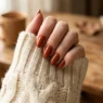 Manicured hand with fall nail design resting on cream knit sweater sleeve in cozy autumn lighting