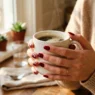 Woman's hands holding white coffee mug showing fresh deep red manicured nails in morning kitchen light