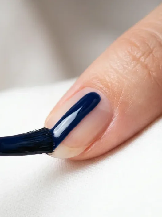 Polish brush applying glossy navy blue lacquer in single stroke down center of fingernail
