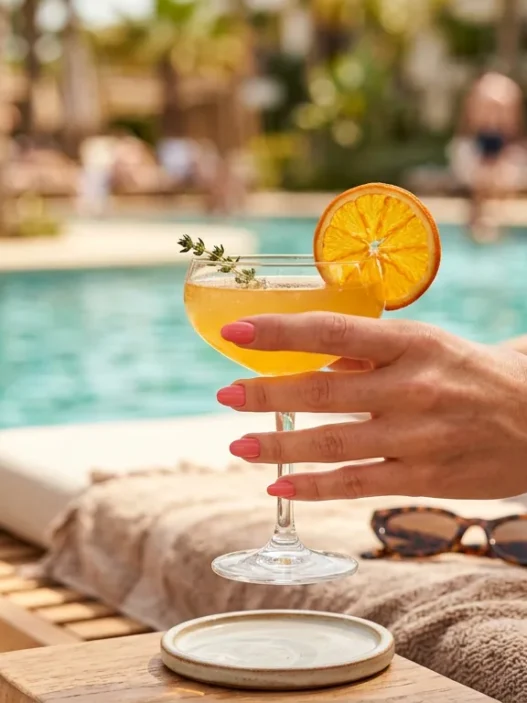 Manicured hand with neutral polish holding citrus cocktail glass on sunny pool deck with turquoise water background