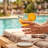 Manicured hand with neutral polish holding citrus cocktail glass on sunny pool deck with turquoise water background
