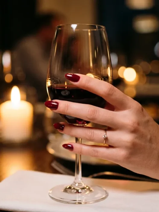 Manicured hand with classic red nails holding wine glass at candlelit dinner table