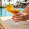Manicured hand with square nails holding cocktail glass by pool with turquoise water bokeh