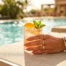 Manicured hand holding cocktail glass with citrus garnish on sunlit pool deck with turquoise water