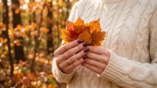 Hands with deep burgundy shimmer nails holding autumn leaves against cream knit sweater in fall sunlight