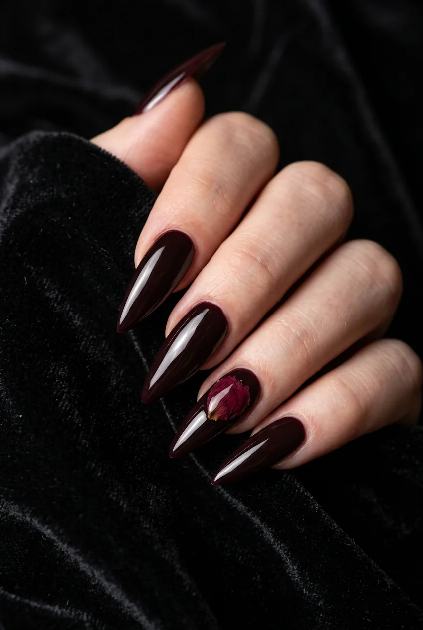 Long stiletto nails in deep black-cherry gel finish photographed against dark velvet with a dried rose petal sealed inside one nail