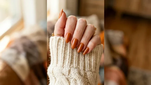 Close-up of hand with burnt caramel satin nails resting on cream chunky knit sweater cuff in warm autumn light