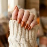 Close-up of hand with burnt caramel satin nails resting on cream chunky knit sweater cuff in warm autumn light