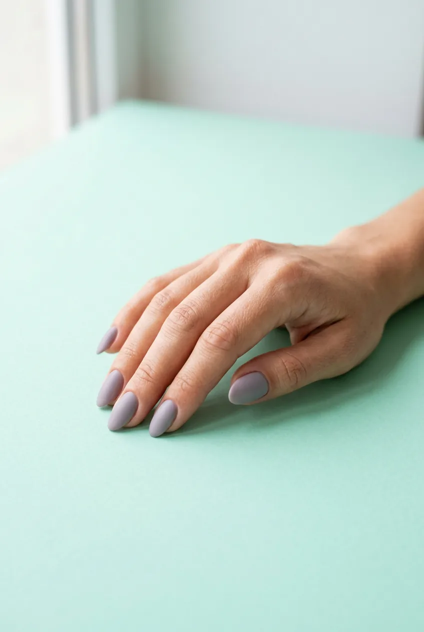 Medium almond nails in dusty grey-toned lilac with a soft matte-cream finish resting on a mint paper backdrop