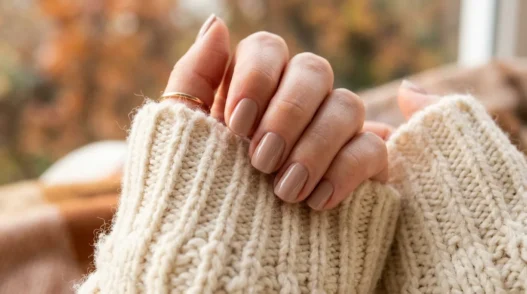 Woman's manicured hand resting on chunky cream knit sweater sleeve in warm cosy autumn light showing fall nail design