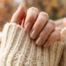 Woman's manicured hand resting on chunky cream knit sweater sleeve in warm cosy autumn light showing fall nail design