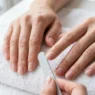 Hands resting on white surface with nails being shaped using a fine-grit nail file, cuticles pushed back and nails bare