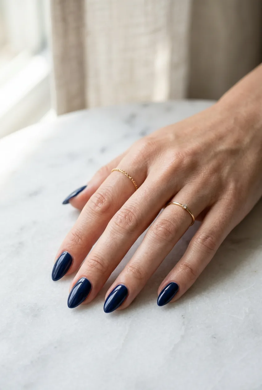 Woman's hand with deep navy blue almond-shaped gel nails resting on white marble in natural window light