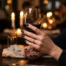 Manicured hand with navy blue gel nails holding a glass of red wine at a candlelit dinner table