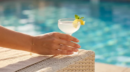 Close-up of a hand with nude blush nails holding a cocktail glass with citrus on a sunny pool deck