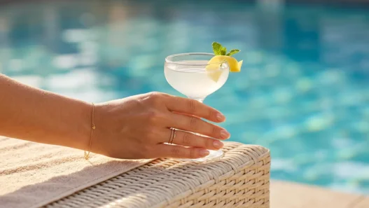 Close-up of a hand with nude blush nails holding a cocktail glass with citrus on a sunny pool deck