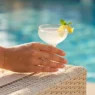 Close-up of a hand with nude blush nails holding a cocktail glass with citrus on a sunny pool deck