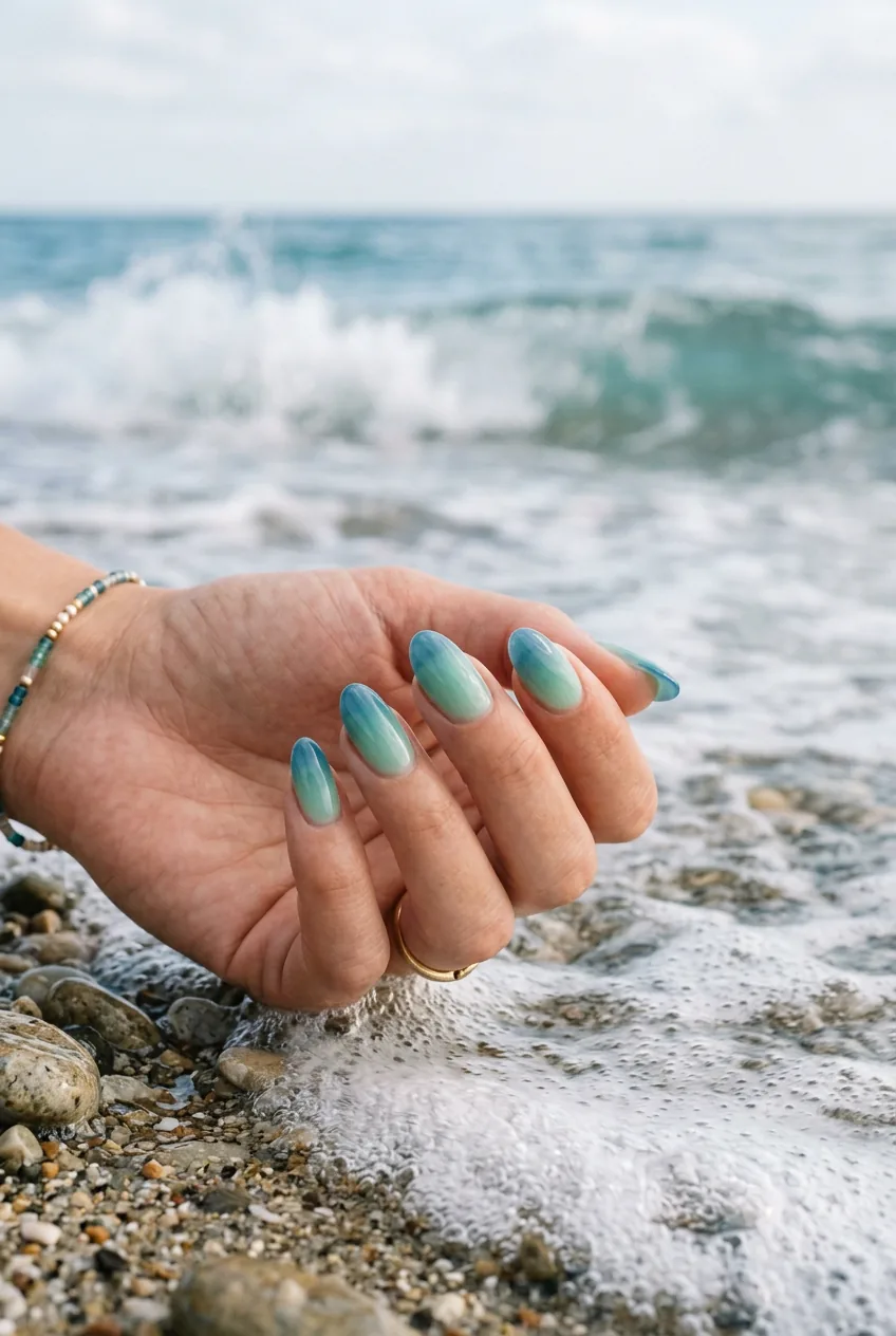 Ombre beach glass nails in blue-green gradient positioned near ocean waves with coastal lighting