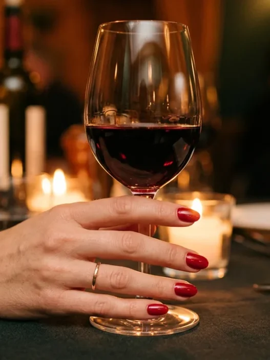 Close-up of a manicured hand holding red wine glass at candlelit restaurant table with warm bokeh lighting
