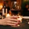 Close-up of a manicured hand holding red wine glass at candlelit restaurant table with warm bokeh lighting