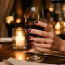 Elegant red manicured hand holding wine glass at romantic candlelit dinner table setting