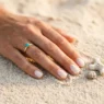 Manicured hand with summer nail design resting on warm beach sand with coral shell beside fingertips