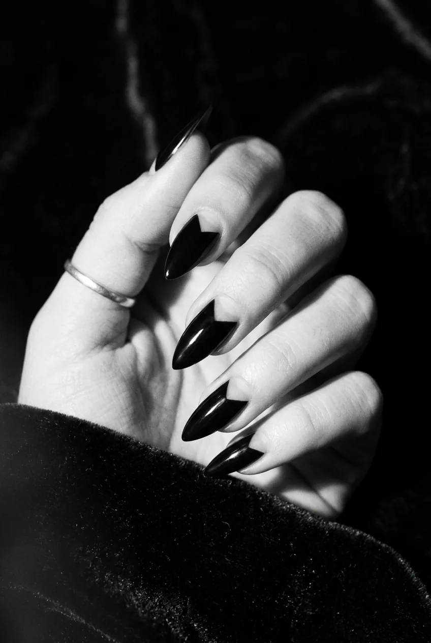 Manicured hand with black stiletto nails featuring geometric negative space cut-outs near cuticle, dramatic split side light on dark background