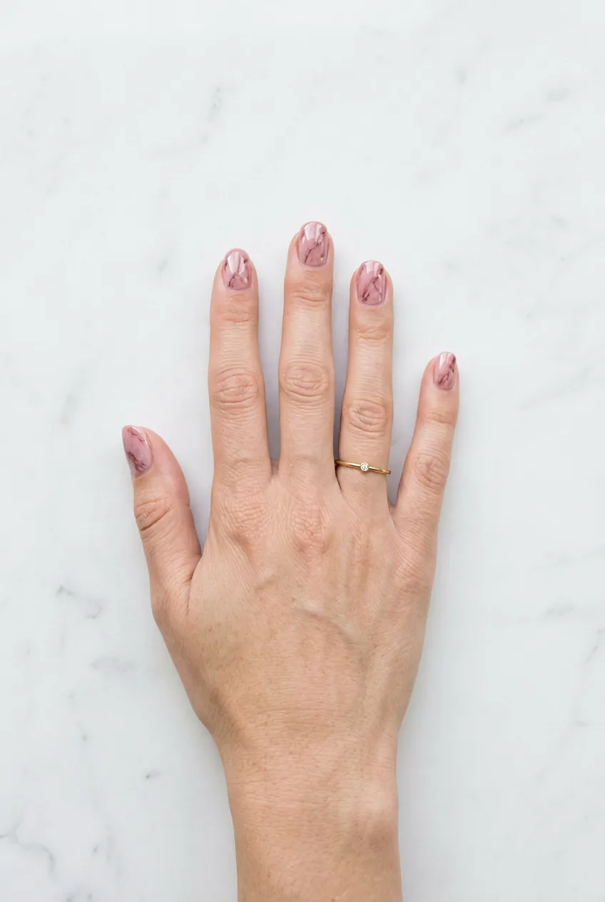 Hand resting on white marble counter with blush rose marble nails and delicate mauve veining alongside a gold ring