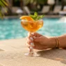 Woman's hand with chrome silver nails holding citrus cocktail glass on sunlit pool deck with turquoise water bokeh