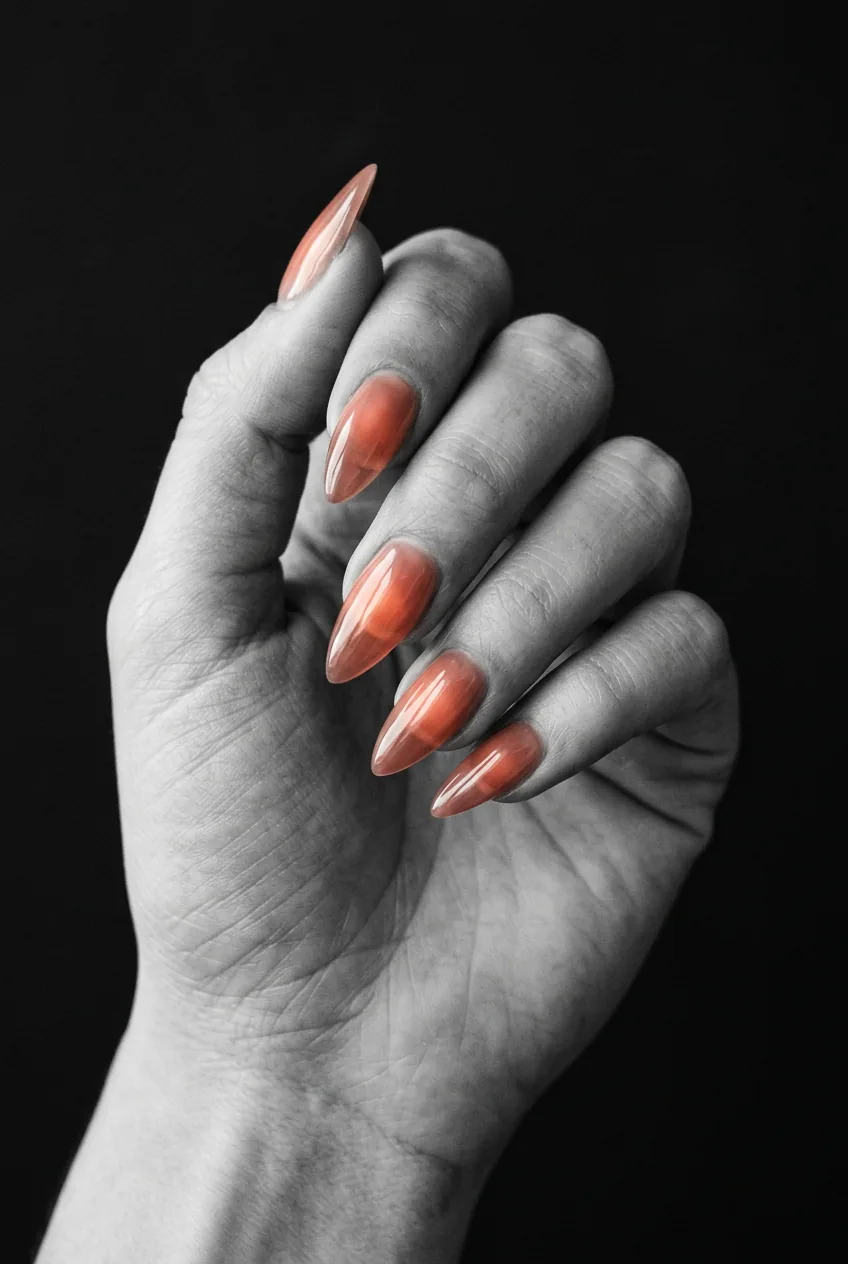 Manicured hand with medium stiletto nails in sheer coral jelly finish glowing under warm directional light on matte black background