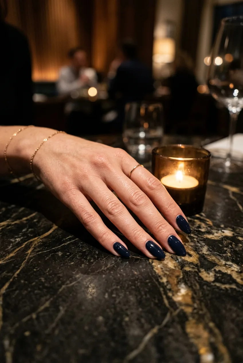 Deep navy near-black almond nails resting on a dark marble restaurant table in warm candlelight