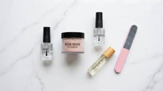 Dip powder nail supplies arranged on a marble surface including base coat, activator, powder jar, nail file, and cuticle oil in natural daylight
