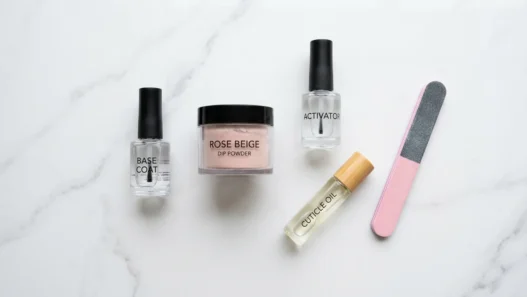 Dip powder nail supplies arranged on a marble surface including base coat, activator, powder jar, nail file, and cuticle oil in natural daylight