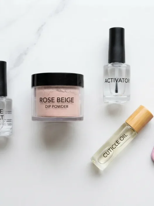Dip powder nail supplies arranged on a marble surface including base coat, activator, powder jar, nail file, and cuticle oil in natural daylight