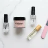 Dip powder nail supplies arranged on a marble surface including base coat, activator, powder jar, nail file, and cuticle oil in natural daylight