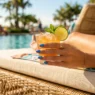 Manicured hand with duochrome blue-violet nails holding a citrus cocktail glass on a sunlit pool deck