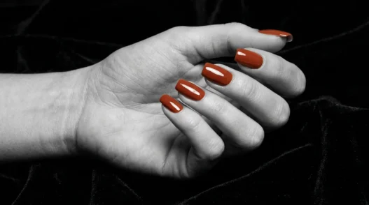 Close-up of manicured nails with a glossy glazed glass finish in warm terracotta against dark velvet background under dramatic side light