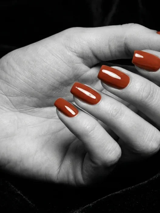 Close-up of manicured nails with a glossy glazed glass finish in warm terracotta against dark velvet background under dramatic side light