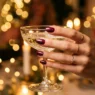 Close-up of manicured hand with deep glossy glitter nails holding champagne coupe in golden party light