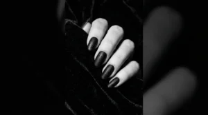 Close-up editorial shot of a manicured hand with deep black Halloween nails against dark velvet background, dramatic side lighting