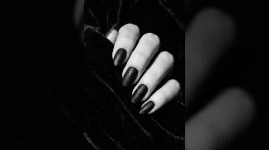 Close-up editorial shot of a manicured hand with deep black Halloween nails against dark velvet background, dramatic side lighting