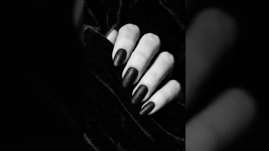 Close-up editorial shot of a manicured hand with deep black Halloween nails against dark velvet background, dramatic side lighting