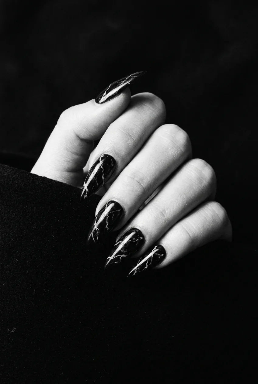 Close-up of long stiletto nails in midnight black marble with white and grey veining running toward sharp tips, dramatic side light