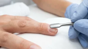 Nail tech pushing back cuticles on a bare natural nail with a metal cuticle pusher under bright salon light